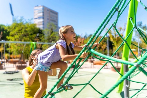 Mother assisting her happy daughter climbing a green rope jungle gym on a sunny day, experiencing