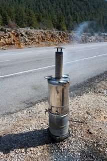 Smoking metal stove with roadside chimney against wooded backdrop, traditional samovar, kettle for