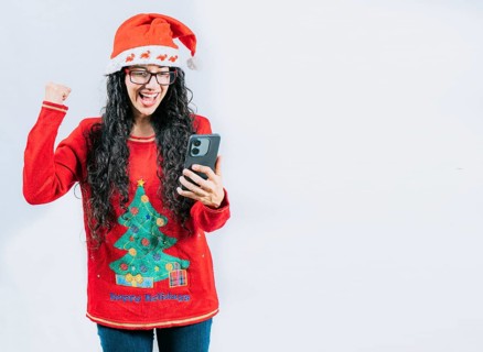 Excited girl in Christmas sweater looking at phone screen, isolated. Young woman in Christmas hat