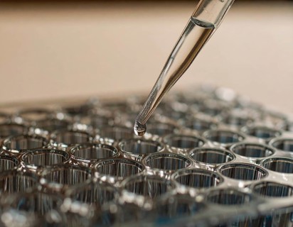 A single droplet hovers at a pipette tip above a microplate bathed in warm, soft light, Pipette