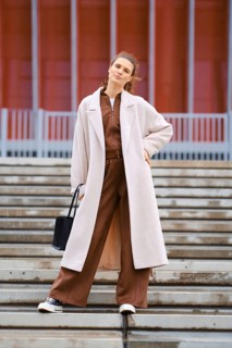 A confident woman stands on the steps wearing a brown tracksuit and a long cream coat. She carries