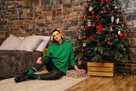 A cheerful woman sits on the floor beside a beautifully decorated Christmas tree in a cozy indoor