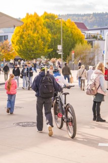 A man pushes his bike across a sidewalk while people and autumn trees form the backdrop,