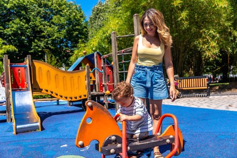 Young mother assisting her son while playing on a rocking horse in a colorful playground on a sunny