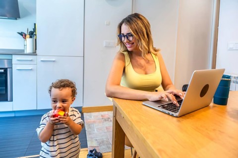Woman working remotely on a laptop in her kitchen while taking care of her baby son eating an apple