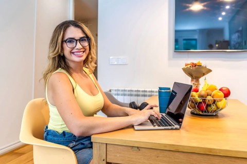 Smiling woman using laptop at kitchen table, working remotely with fruit basket and headphones
