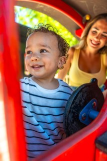 Smiling toddler playing joyfully on a playground slide while mother watches nearby, creating