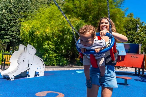 Young mother having fun with her baby son, pushing him on a swing in a colorful playground during a
