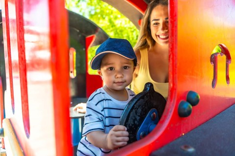 Happy toddler having fun driving a toy train at the playground while his mother watches him with a