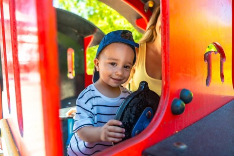Little boy having fun driving a toy train at the playground, with his mother supervising in the