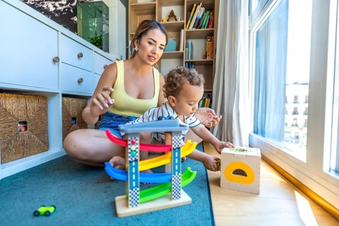 Young mother watching her baby son playing with a toy car garage and a shape sorter on the floor at