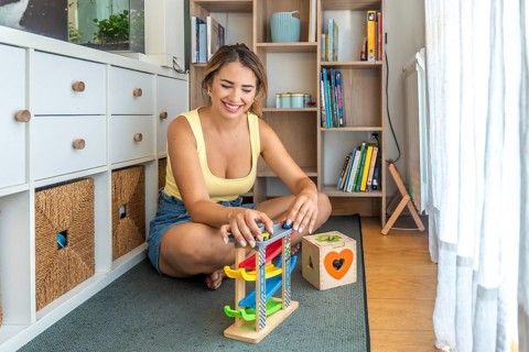 Young woman playing with a toy car ramp and smiling while sitting on a play mat in a child's