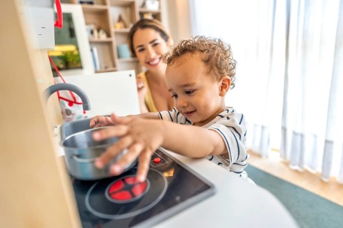 Happy toddler boy having fun playing with a toy kitchen while his mother watches over him in a