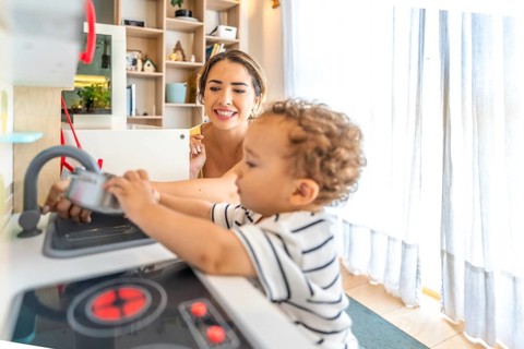 Smiling mother looking at her little son having fun playing with a toy kitchen in a modern, bright