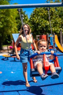 Young child enjoying a swing at the playground with their mother looking on, having fun on a summer