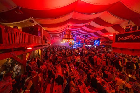 Full beer tent with people, red ceiling and stage, lively and festive atmosphere, Volksfest