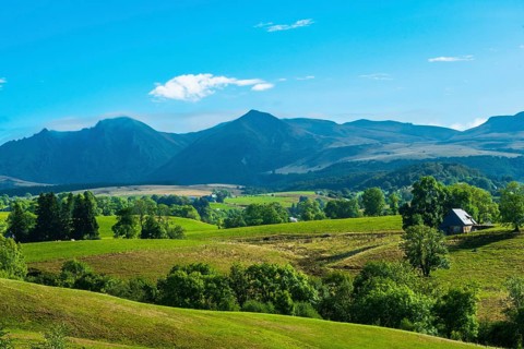 Lush green fields with a solitary house, set against majestic mountains and a bright sky, Massif of