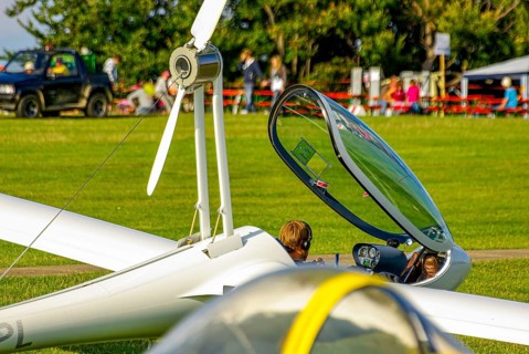 A motorised glider parked at the edge of the airfield with the canopy open during an air show at