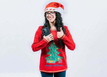 Smiling girl in Christmas hat holding phone with thumb up isolated. Positive young woman in