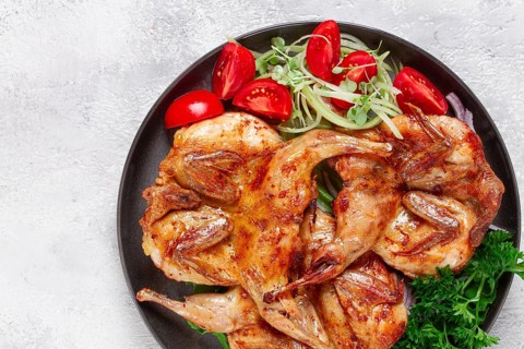 Baked quail, served on a plate with crispy lettuce leaves, fresh tomatoes and herbs, on a dark