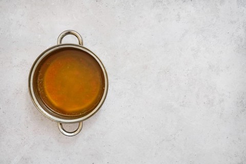 A silver pot filled with rich, golden mushroom broth on a smooth light gray surface. The broth