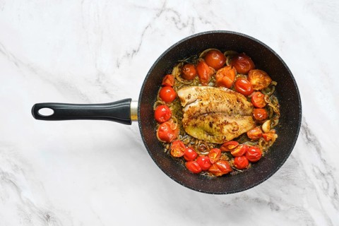 A frying pan holds a seasoned fish fillet surrounded by bright cherry tomatoes. The scene captures