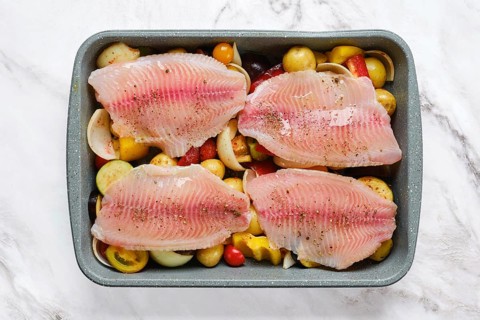Fresh white fish fillets are arranged on a bed of colorful vegetables, including tomatoes and bell