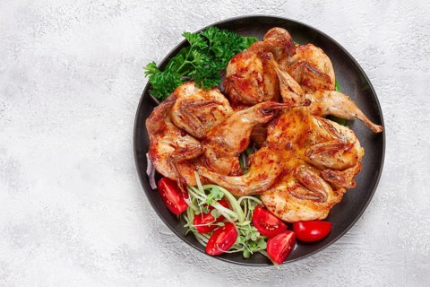 Baked quail, served on a plate with crispy lettuce leaves, fresh tomatoes and herbs, on a dark