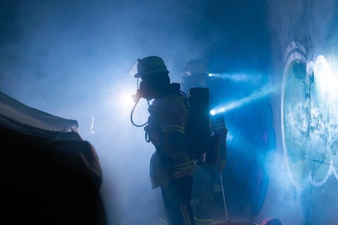 Firefighters in protective suits work in the fog, beams of light cut through the smoke,