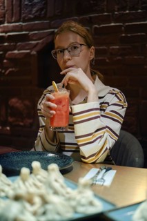 A young woman sits at a wooden table in a warm, dimly lit restaurant. She holds a colorful drink