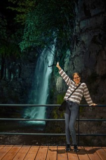 A cheerful young woman stands on a wooden walkway behind a railing, posing joyfully with one arm