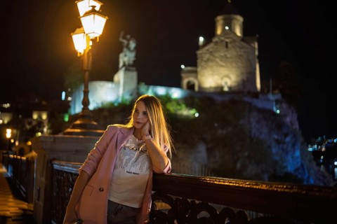 A woman leans against a railing at night, dressed casually with a light jacket. Behind her, a