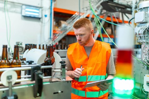 Young man in safety vest checking beer bottles on a factory conveyor belt, performing quality