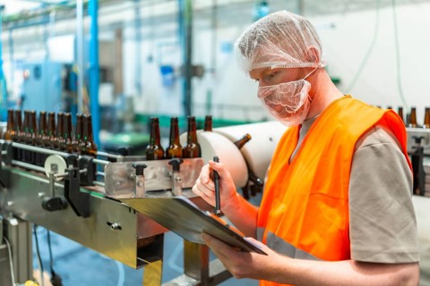 Worker in a hairnet and face mask checking clipboard records while brown beer bottles move along a
