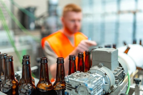 Brown glass bottles moving along a conveyor belt, with a factory worker in an orange vest