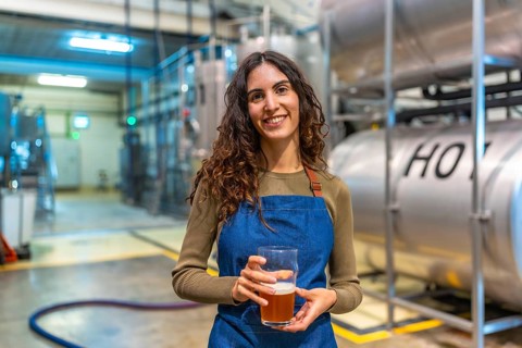 Woman brewer standing in modern brewery, smiling and holding a glass of craft beer for quality