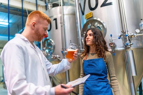 Brewery workers evaluating beer sample for quality control, holding clipboard and glass in brewing