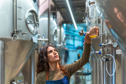 Woman brewer performing quality control, holding a glass sample of fresh beer, standing in a modern