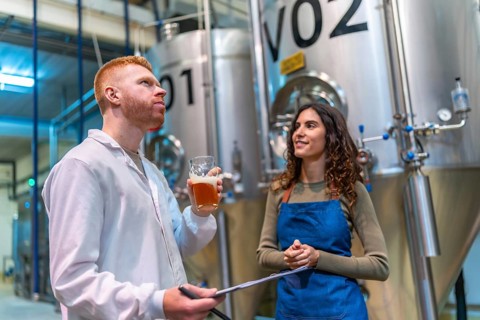 Two brewery professionals inspect and taste craft beer beside large fermentation tanks in a modern