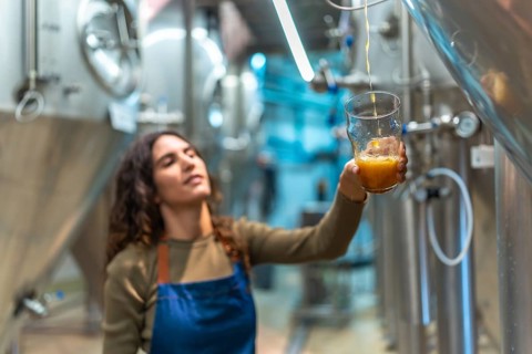 Female brewer wearing an apron, carefully pouring freshly brewed craft beer into a glass for a