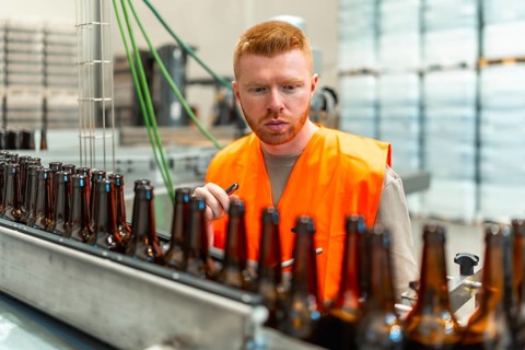 Man wearing an orange safety vest inspecting empty glass beer bottles moving along a conveyor belt
