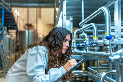 Woman wearing a lab coat meticulously checking industrial pipes and valves in a modern beverage