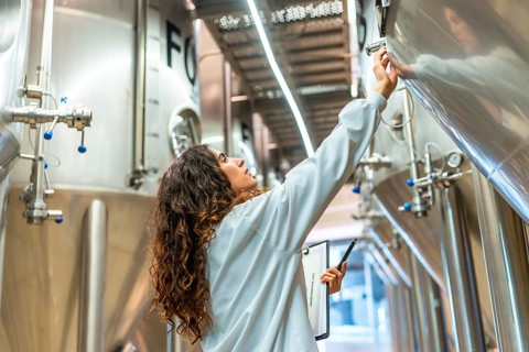 Young woman in a lab coat checking control panel on a stainless steel fermentation tank, performing