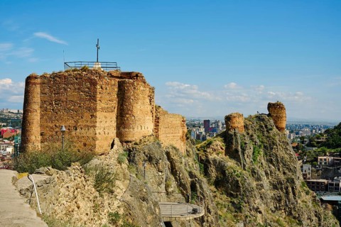 A weathered stone fort stands on a rocky hill, crowned with a cross. Below, a vibrant city