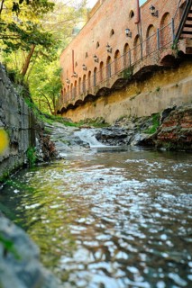 A clear stream flows calmly past old brick structures surrounded by lush greenery. Sunlight