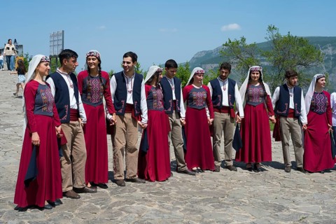 Group in red traditional costumes dancing together on cobblestones against a mountain backdrop,
