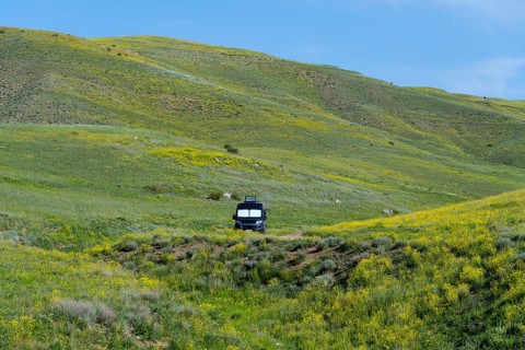 A black van stands alone in a meadow next to green and yellow hills under a blue sky, camper,