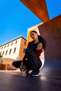 Young woman squatting on an urban surface, posing casually against a modern rust colored