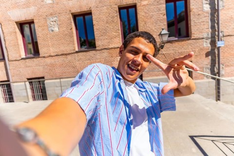 Young man smiling and making a playful hand gesture while taking a selfie in sunny urban streets,