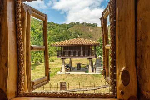Open wooden window with rope hinges framing a scenic view of a traditional elevated horreo granary